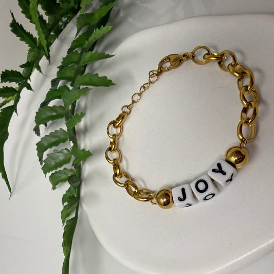 A gold-tone chain bracelet with three white cylindrical beads printed with the letters 'J', 'O', and 'Y', displayed on a white ceramic dish with green fern leaves in the background.